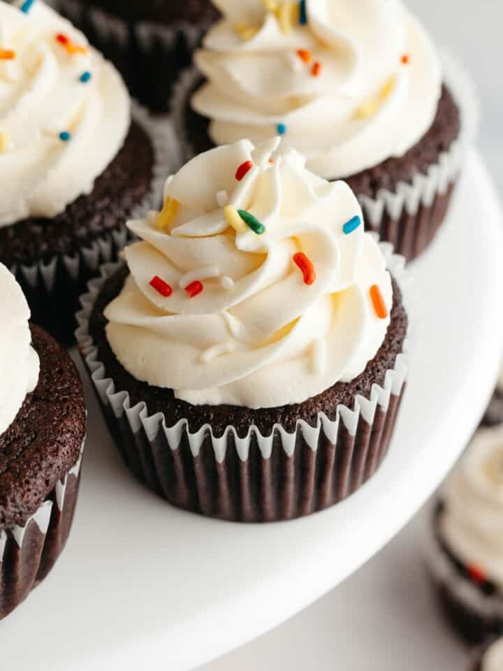 Chocolate cupcakes with a white frosting and rainbow sprinkles on a cake stand.