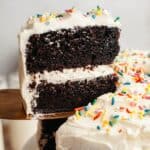 A slice of chocolate cake with vanilla frosting being lifted up with a cake server.
