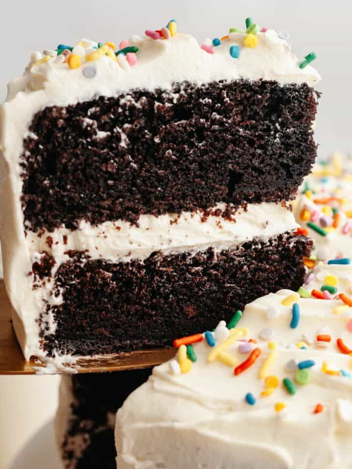 A slice of chocolate cake with vanilla frosting being lifted up with a cake server.