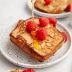 Two slices of buttermilk french toast on a small plate with raspberries with maple syrup being poured on top.