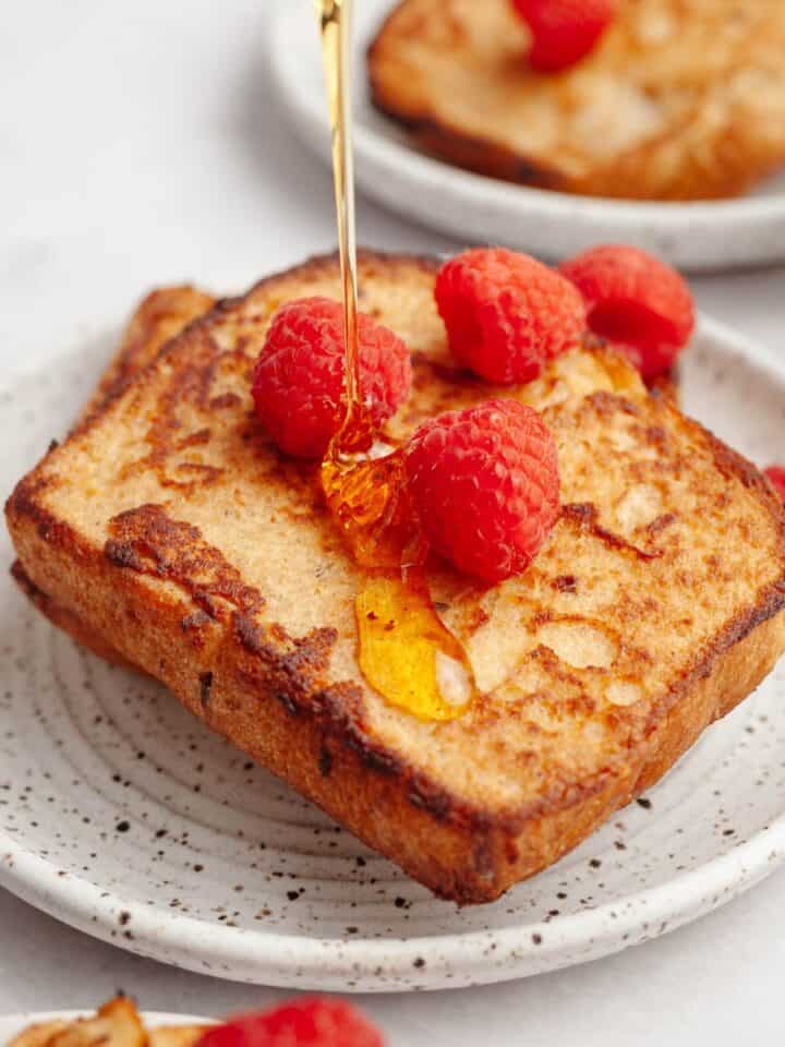 Two slices of buttermilk french toast on a small plate with raspberries with maple syrup being poured on top.