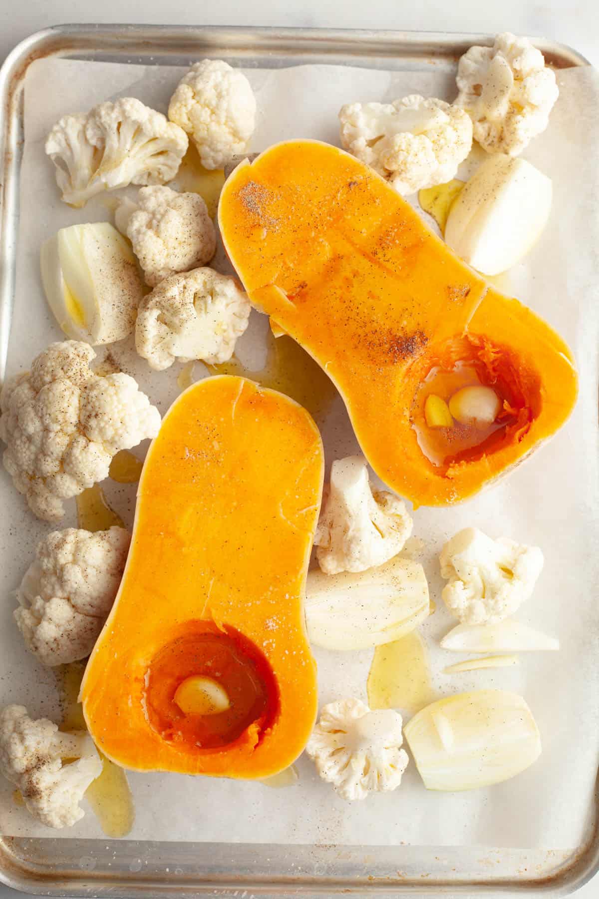 A sheet pan with a butternut squash cut in half, chopped onions and cauliflower florets with olive oil and pepper.