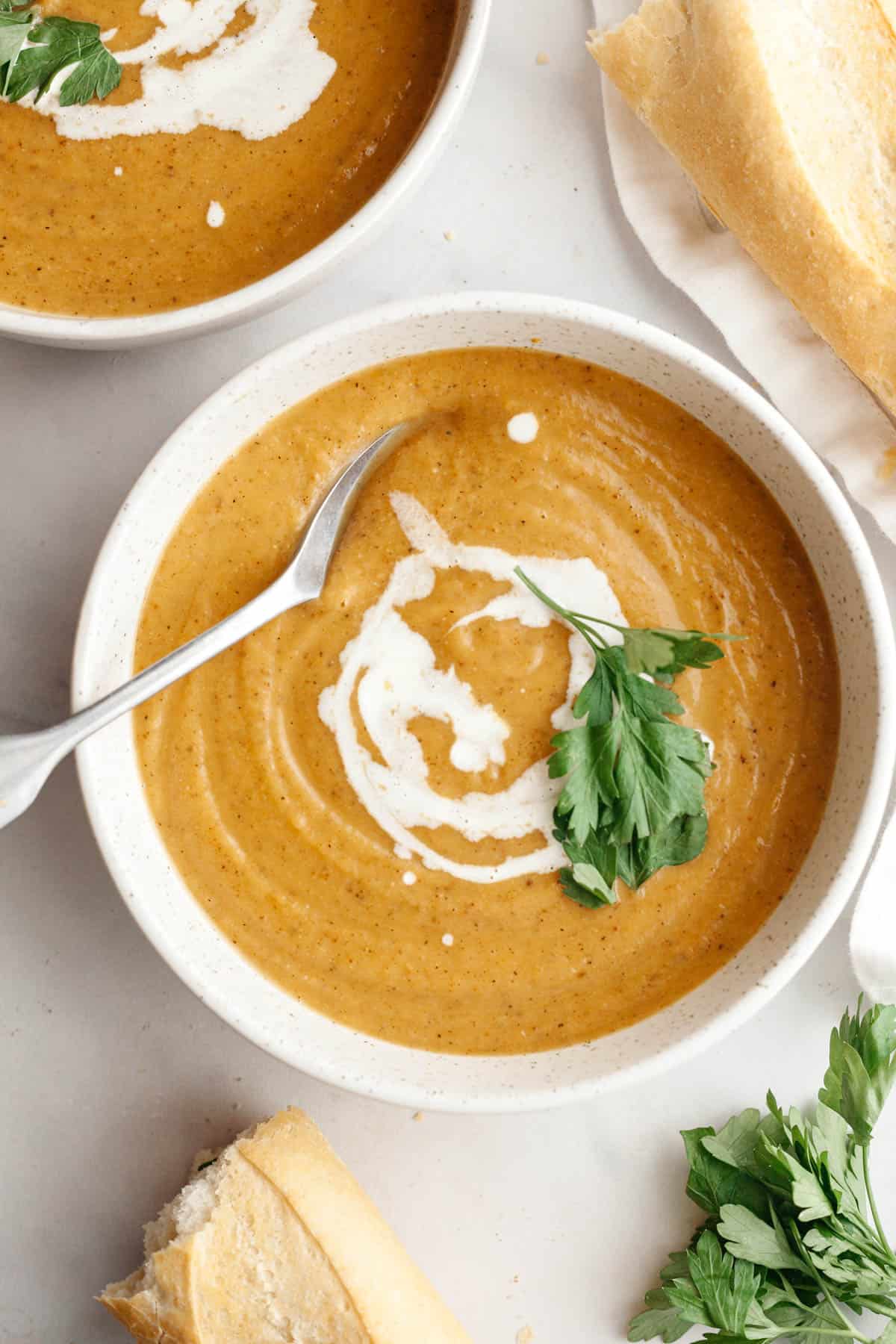 A white soup bowl with butternut squash cauliflower soup, drizzled with cream and garnished with herbs, there is a spoon in the soup.