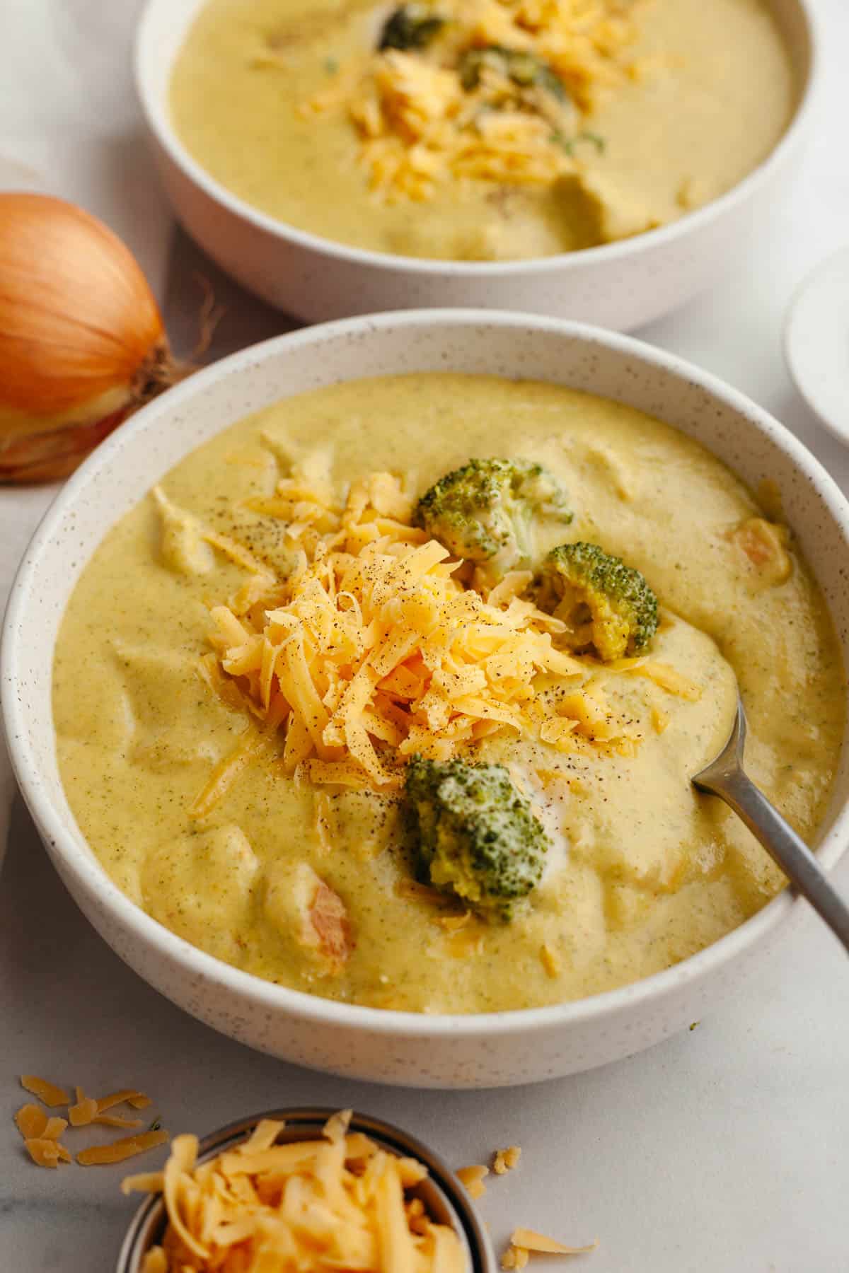 A bowl of chicken and broccoli cheddar soup, topped with shredded cheddar cheese.