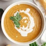A white soup bowl with butternut squash cauliflower soup, drizzled with cream and garnished with herbs.