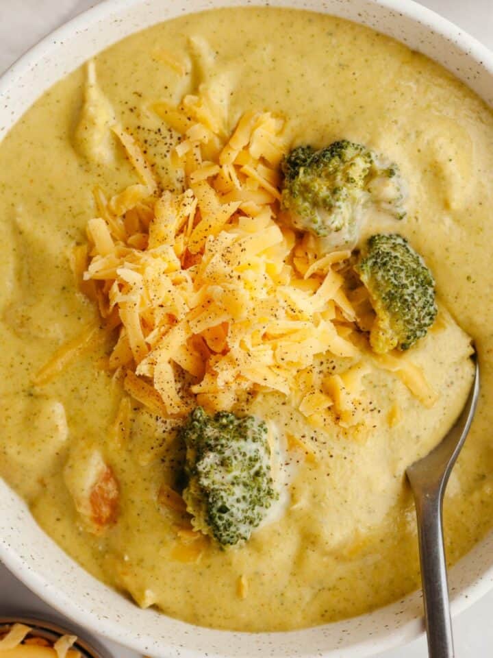 A bowl of chicken and broccoli cheddar soup, topped with shredded cheddar cheese.
