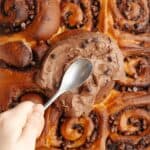 A hand spread chocolate cream cheese frosting with a spoon on top of chocolate cinnamon rolls.