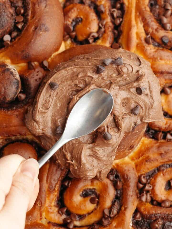 A hand spread chocolate cream cheese frosting with a spoon on top of chocolate cinnamon rolls.