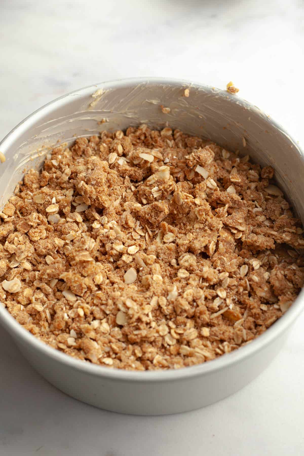 A round cake pan with an unbaked cardamom almond cake covered in an oaty streusel.