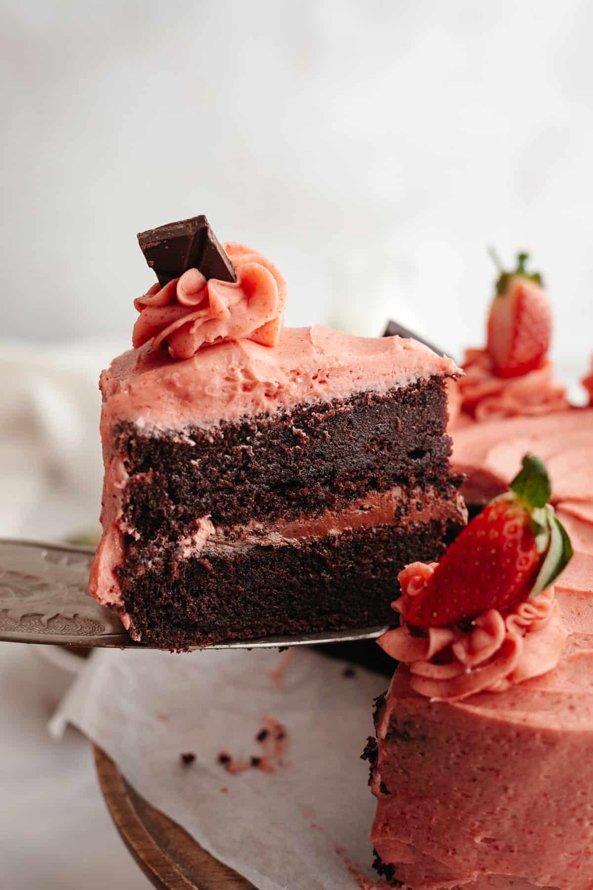 A slice of chocolate cake covered in strawberry frosting being lifted out with a cake server.