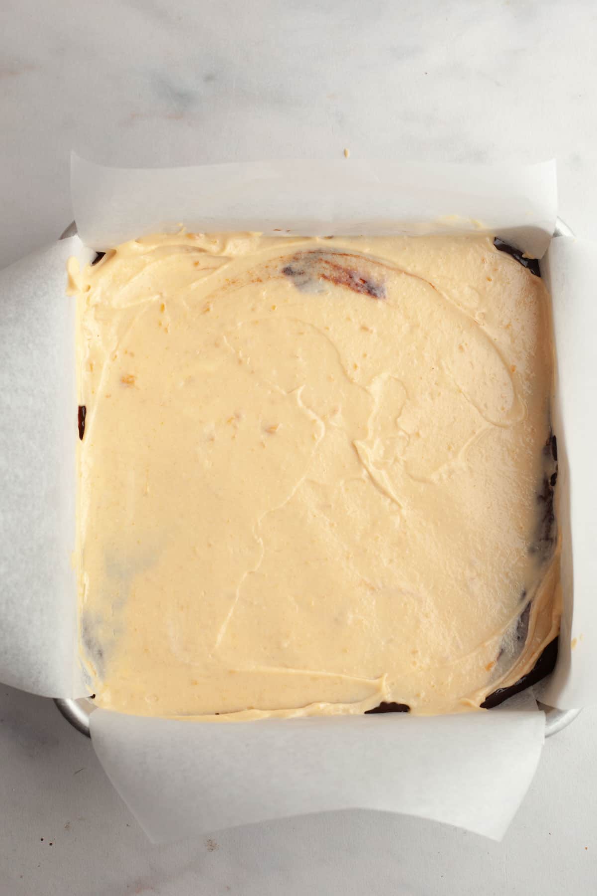 A square pan lined with parchment paper with brownies and peanut butter cheesecake batter on top.