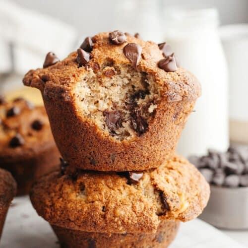 Two jumbo banana muffins with chocolate chips stacked on top of each other, one has a bite taken out of it.