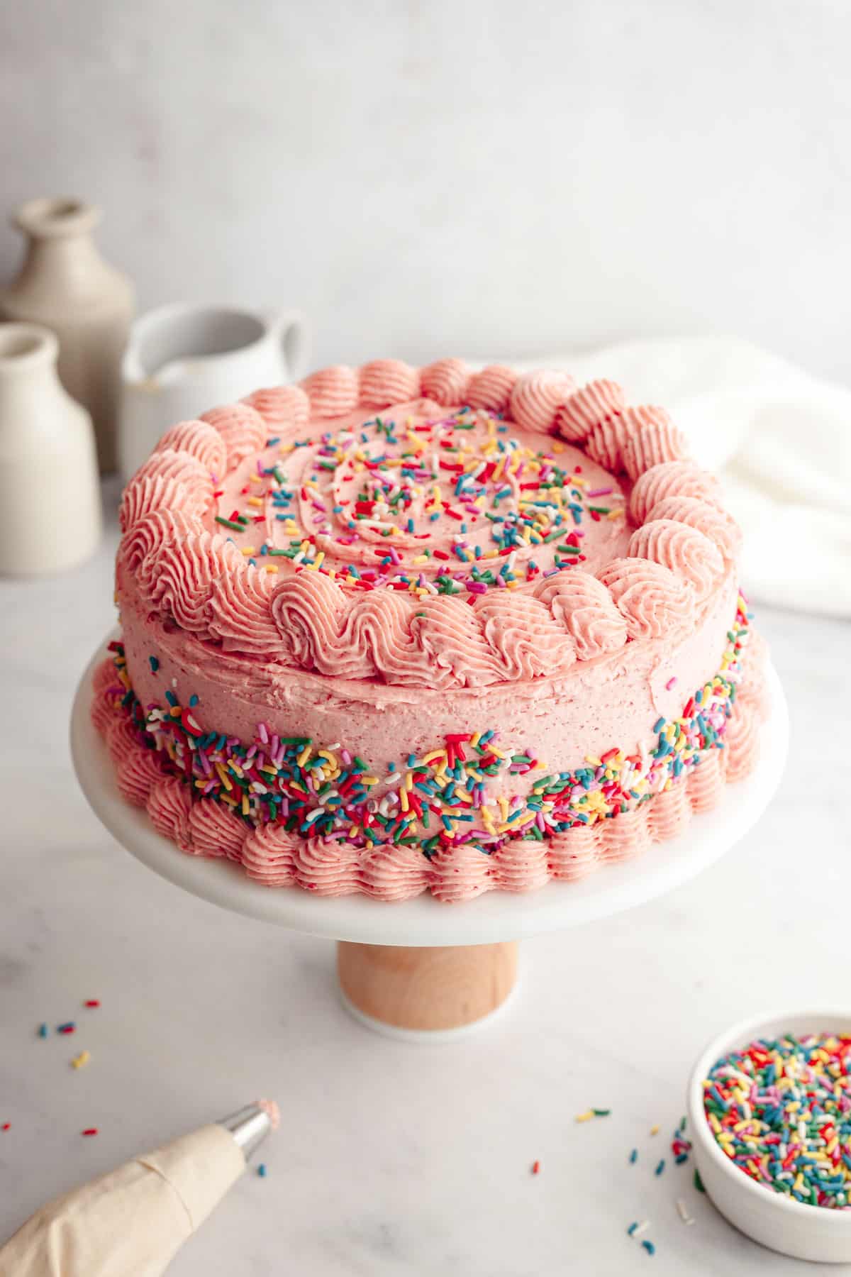 A strawberry frosted cake decorated with sprinkles on a white cake stand.