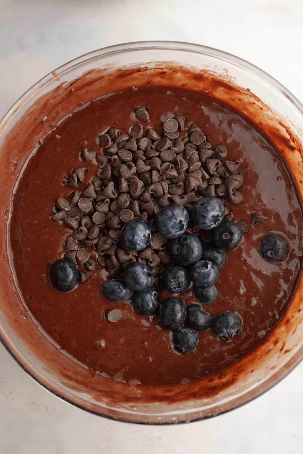 A glass bowl with chocolate muffin batter, chocolate chips and blueberries.