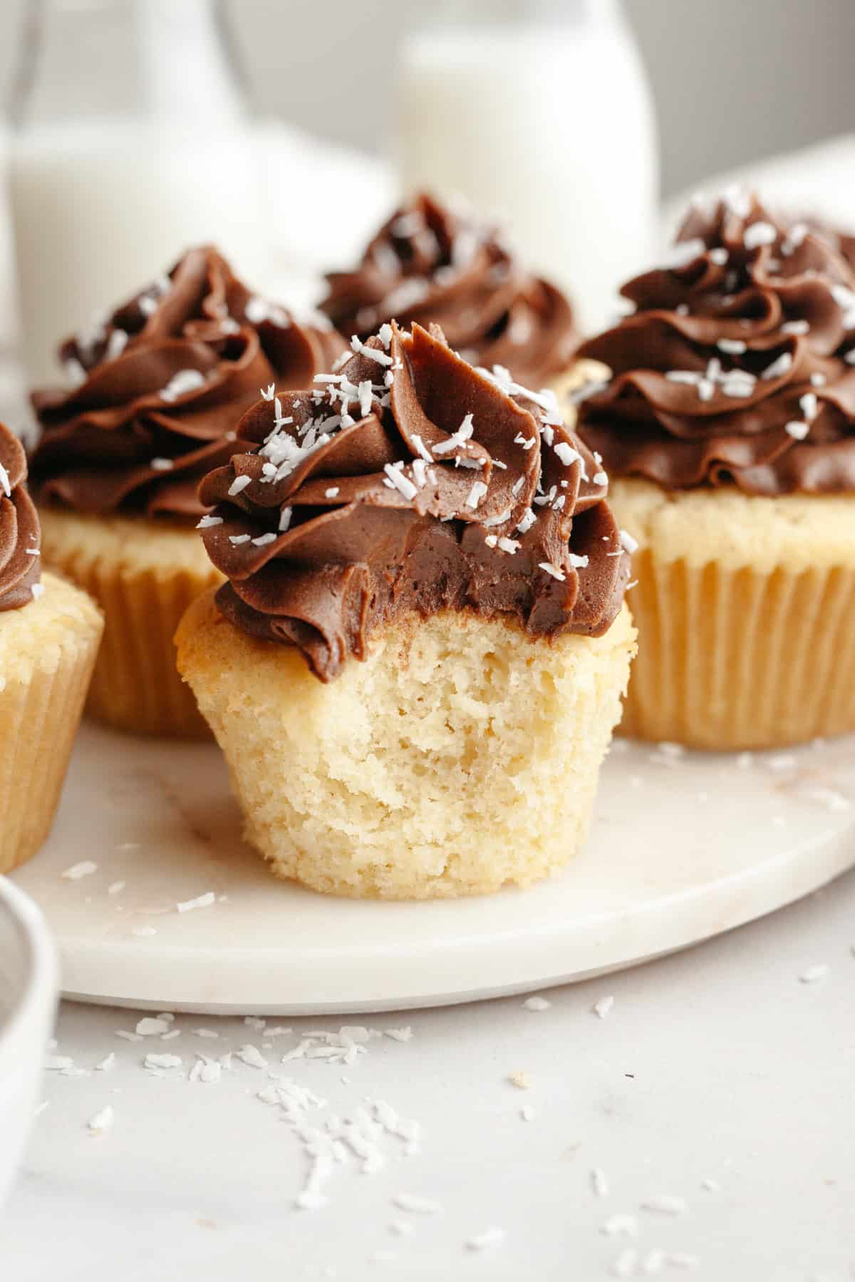 Coconut cupcakes with chocolate frosting, sprinkled with shredded coconut. One cupcake has a bite taken out of it.
