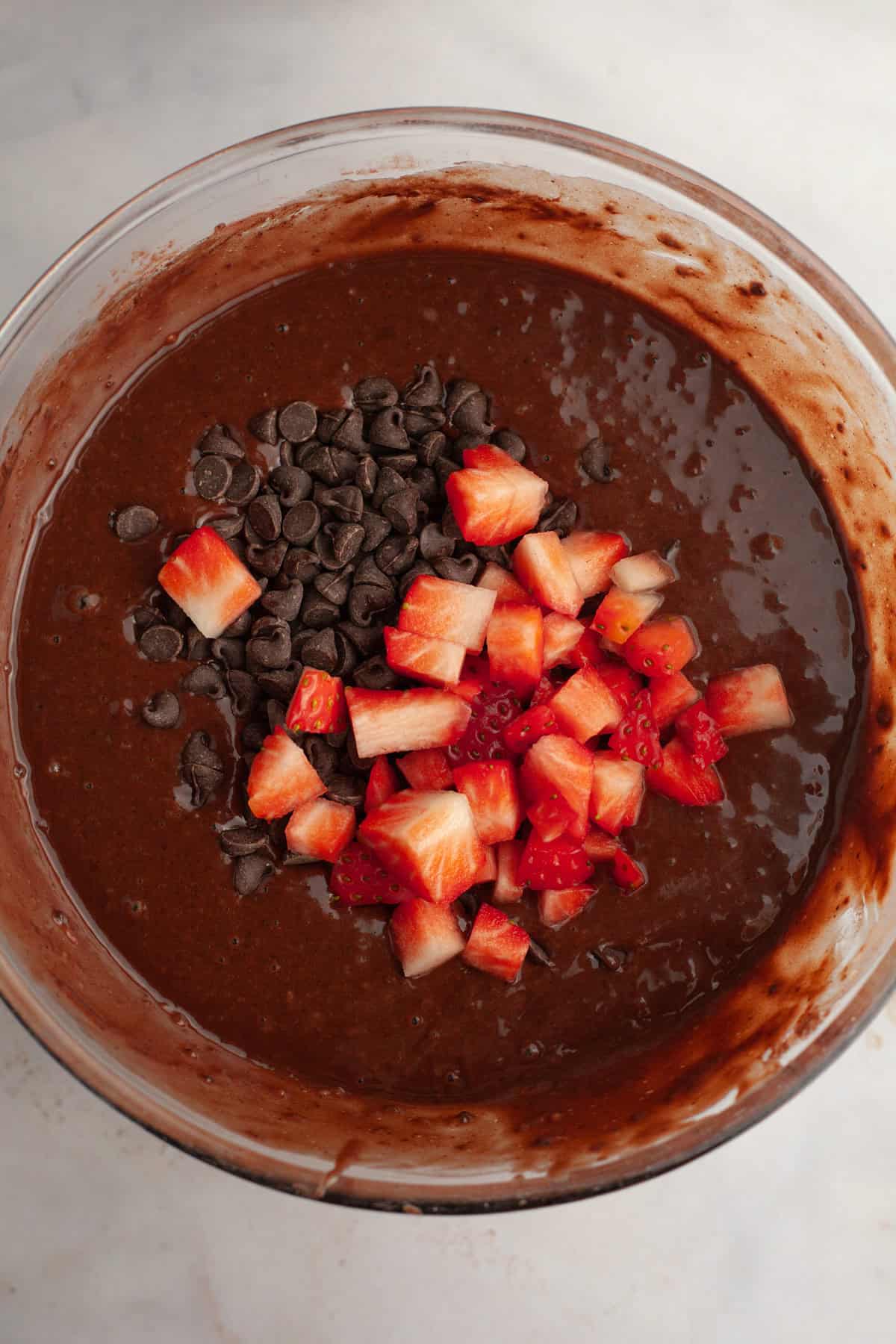 Chocolate muffin batter in a bowl with chocolate chips and chopped up strawberries.