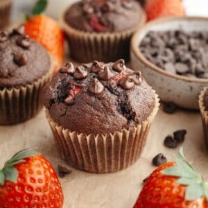 A close up of a chocolate strawberry muffin on parchment paper next to some fresh strawberries.