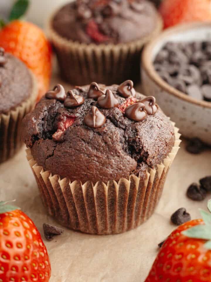 A close up of a chocolate strawberry muffin on parchment paper next to some fresh strawberries.
