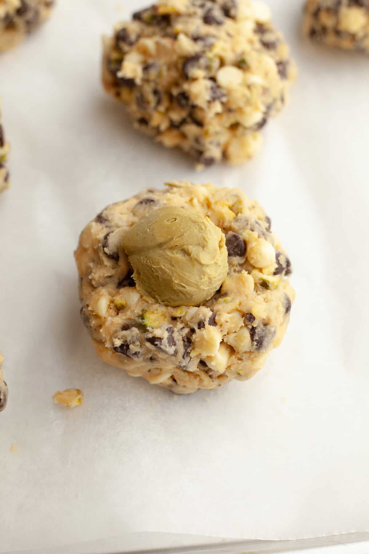 Pistachio chocolate chip cookie dough balls on parchment paper, one has a pistachio butter ball on top.