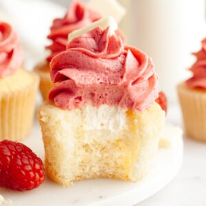A vanilla cupcake with a bite taken out of it, it has a white chocolate ganache filling and pink frosting.