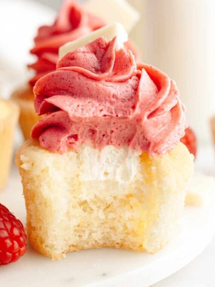 A vanilla cupcake with a bite taken out of it, it has a white chocolate ganache filling and pink frosting.