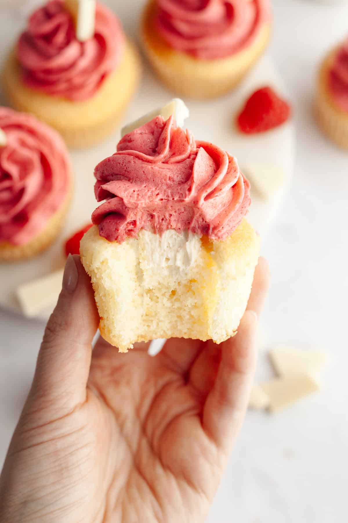 A hand holding a vanilla cupcake with a bite taken out of it, it has a white chocolate ganache filling and pink frosting.