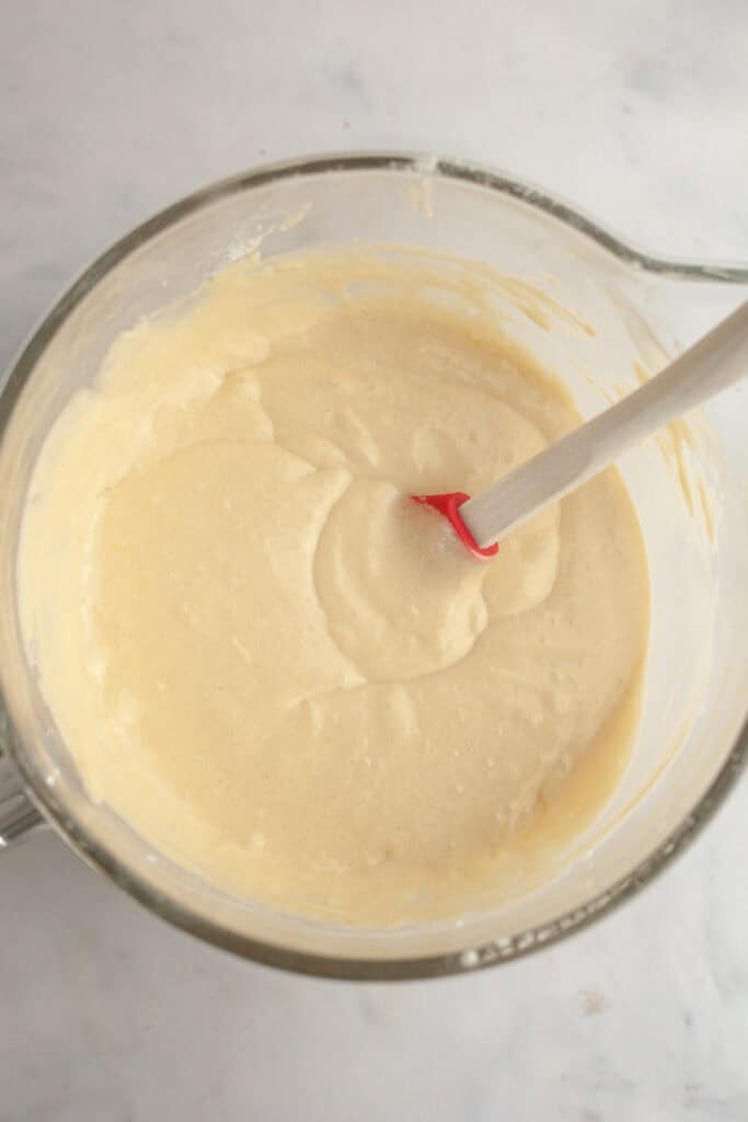 Glass mixing bowl filled with pale cake batter, with a red and white spatula resting inside on a light countertop.