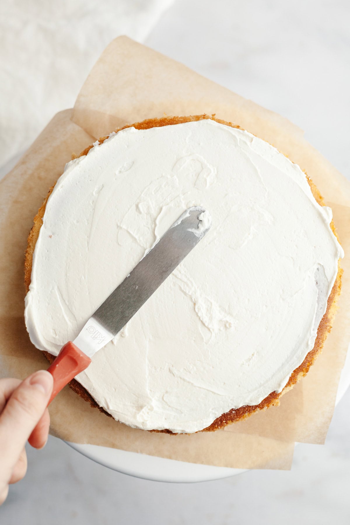 A vanilla cake layer is on parchment paper, a hand is spread over whipped mascarpone frosting with a small offset spatula.