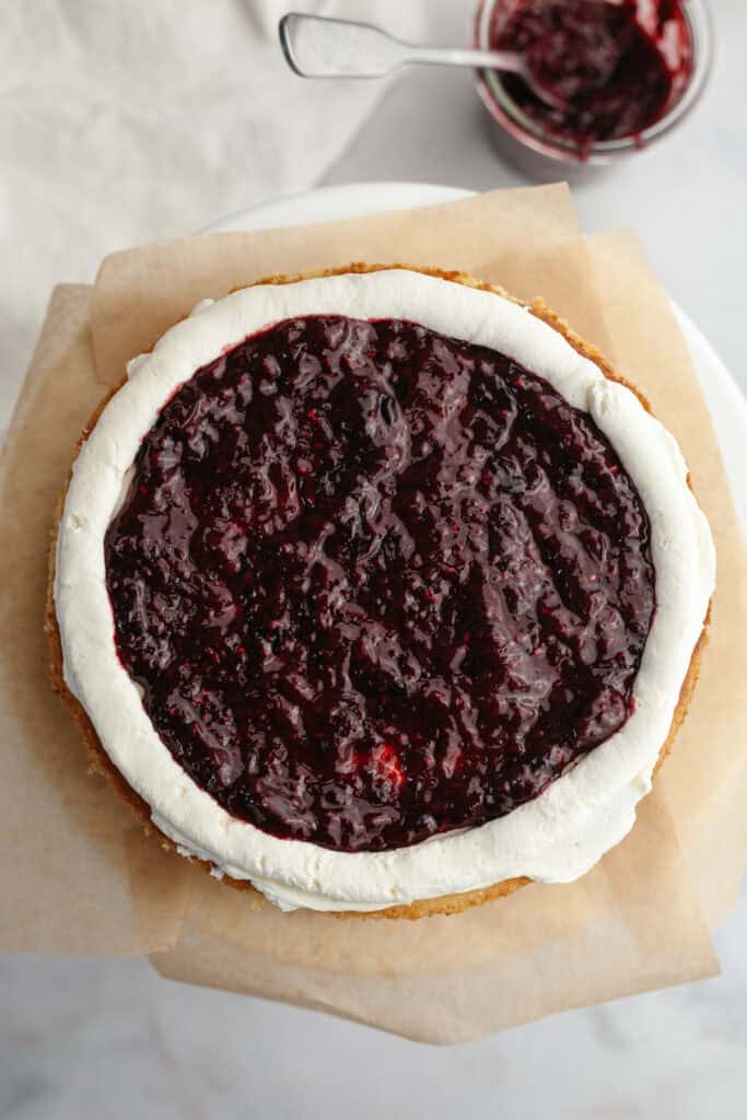 A vanilla cake is on brown parchment paper, it has a ring of white mascarpone frosting around the edges and the middle is covered with a mixed berry compote.