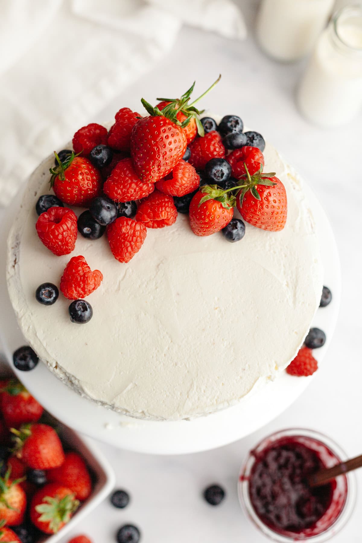 A round berry vanilla cake with white frosting, topped with fresh strawberries, raspberries, and blueberries, sits on a white surface next to a bowl of berries and a jar of jam.