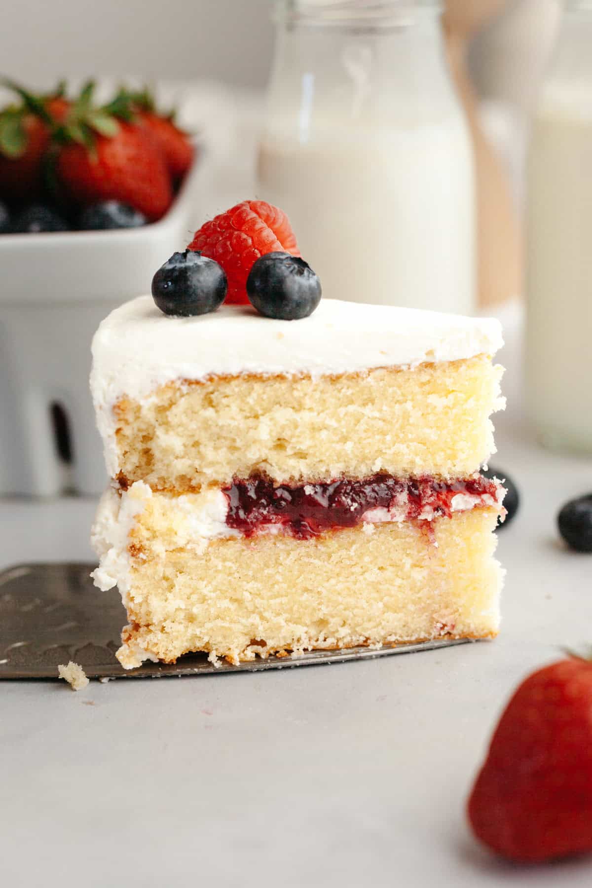 A slice of vanilla sponge cake with white frosting, berry jam filling, and topped with blueberries and a raspberry.