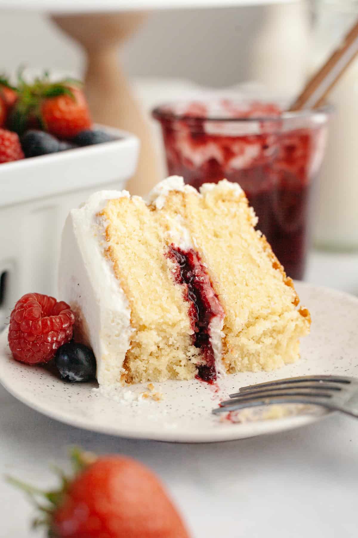 A slice of a berry vanilla layer cake with white frosting, berry jam filling, and topped with a raspberry and blueberry, on a plate with strawberries and jam jar in the background.