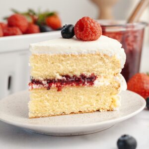 A slice of a berry vanilla layer cake with white frosting, berry jam filling, and topped with a raspberry and blueberry, on a plate with strawberries and jam jar in the background.