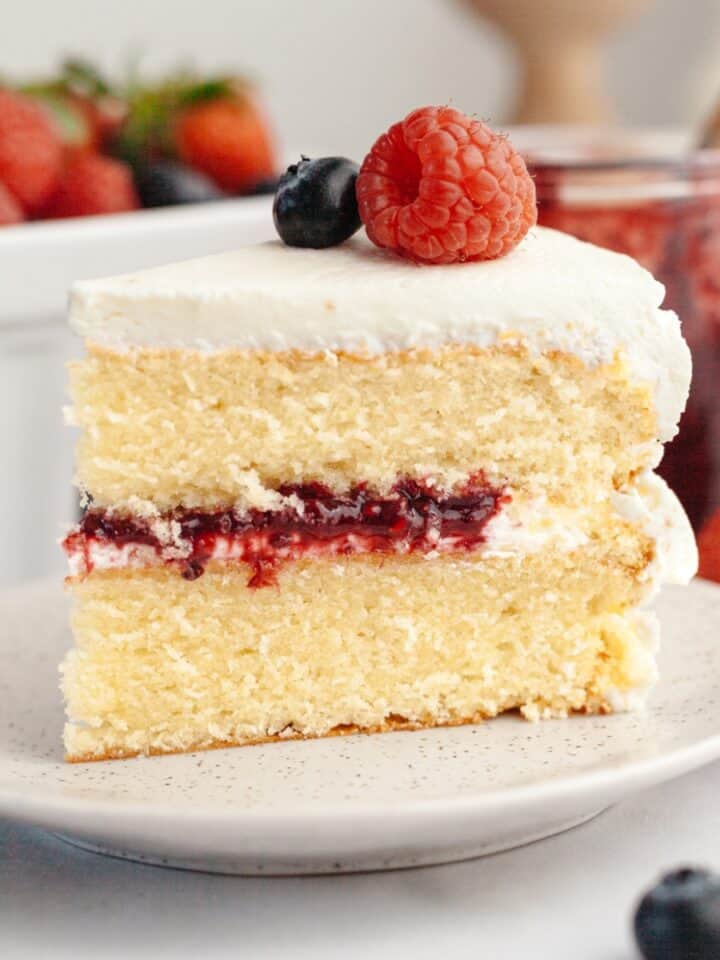 A slice of a berry vanilla layer cake with white frosting, berry jam filling, and topped with a raspberry and blueberry, on a plate with strawberries and jam jar in the background.