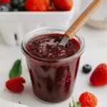Mixed berry compote in a glass jam jar with a wooden spoon. There are fresh berries and mint leaves in the background.