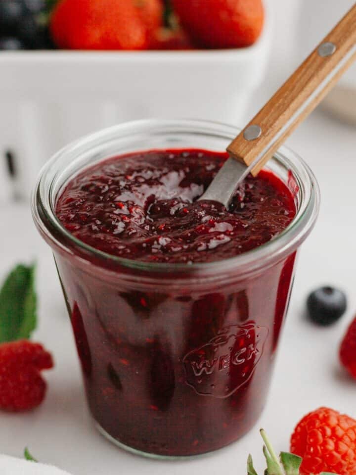Mixed berry compote in a glass jam jar with a wooden spoon. There are fresh berries and mint leaves in the background.