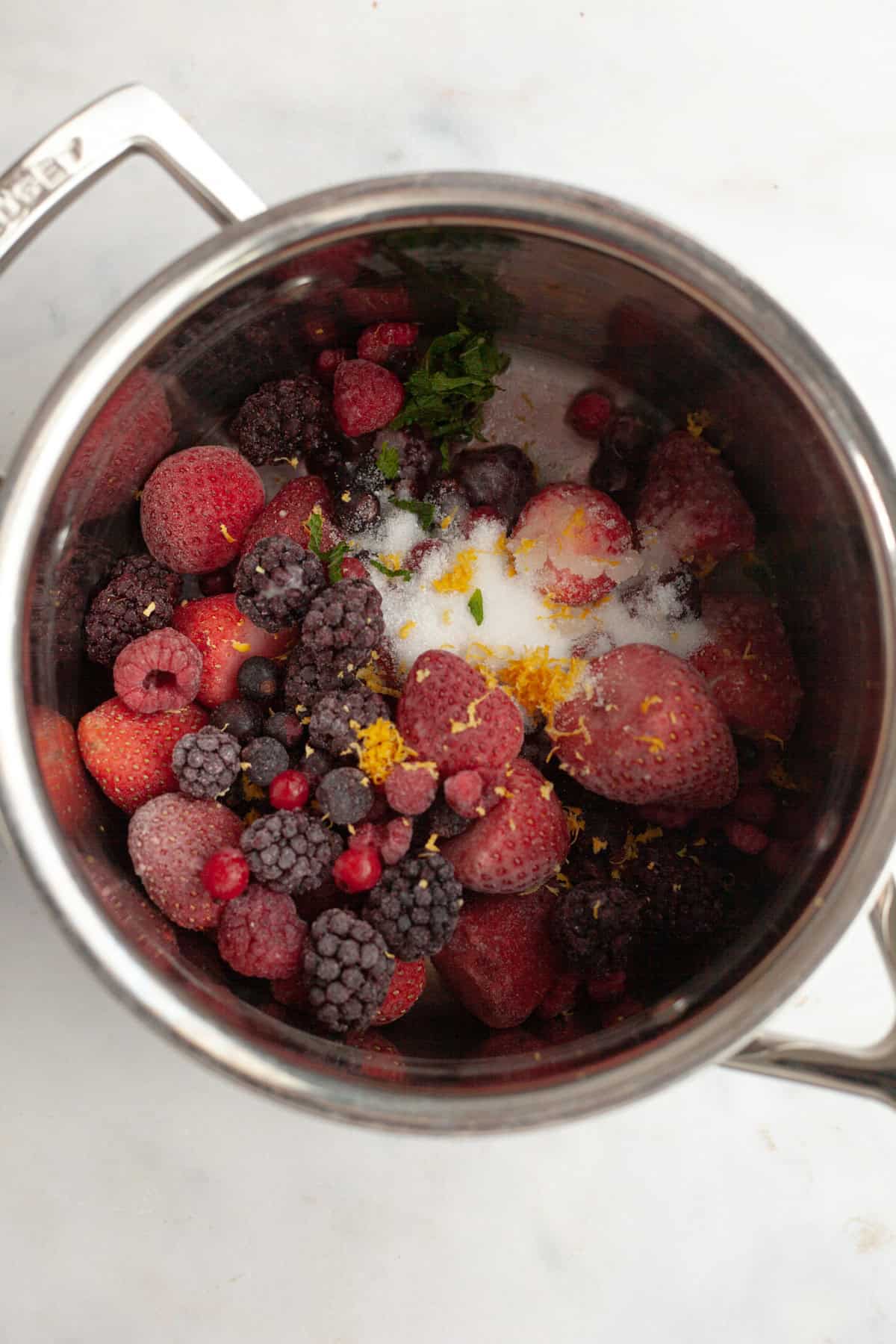 A silver saucepan filled with frozen mixed berries, lemon zest, and sliced up mint leaves.