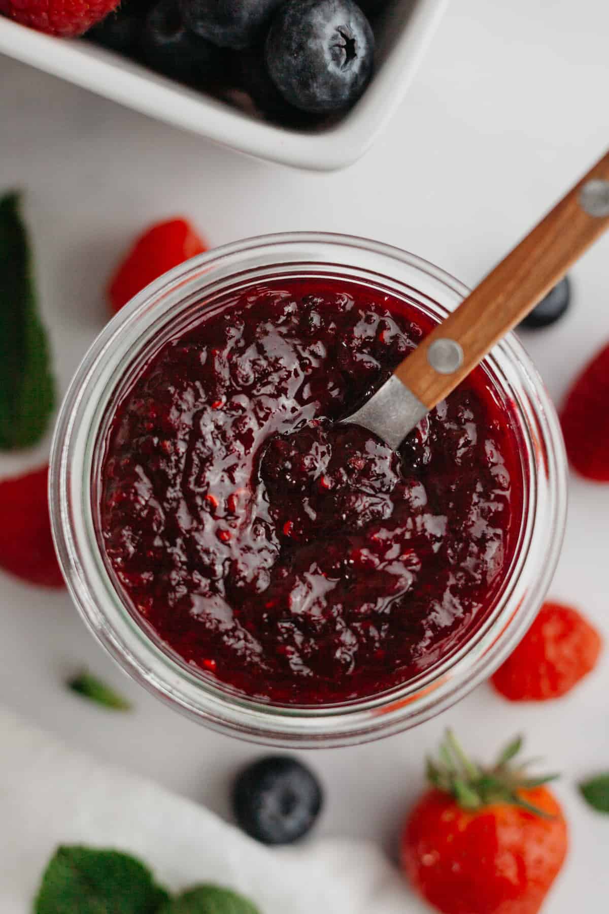 Mixed berry compote in a glass jam jar with a wooden spoon. There are fresh berries in the background.