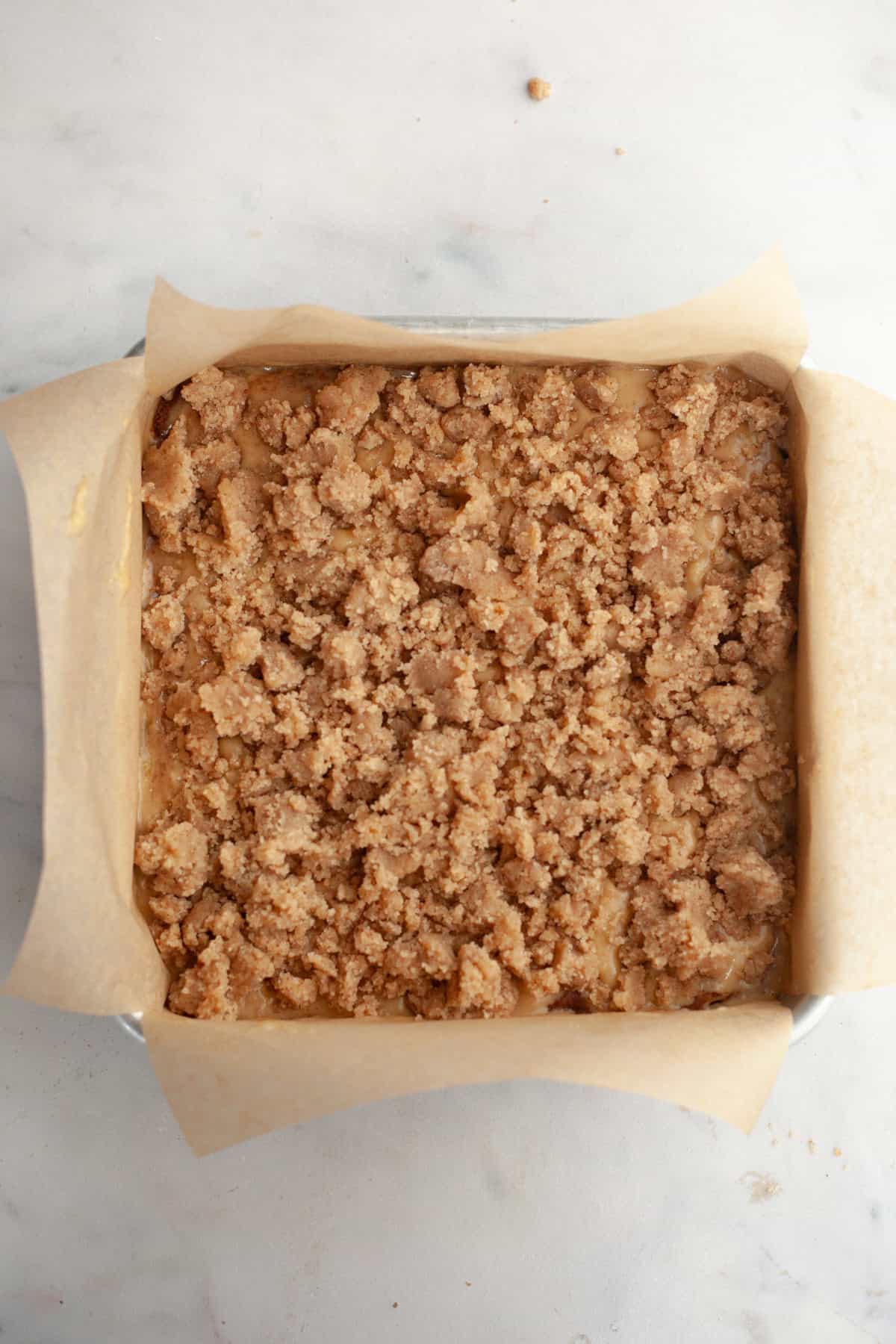 A square baking pan lined with parchment paper and filled with cake batter, topped with streusel.