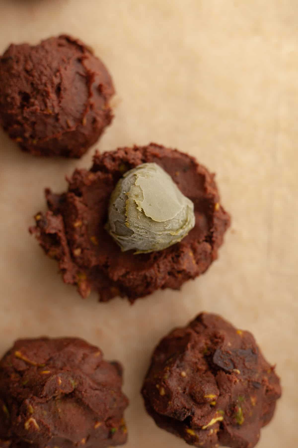 Chocolate cookie dough balls on parchment paper, one is being filled with a ball of pistachio spread.