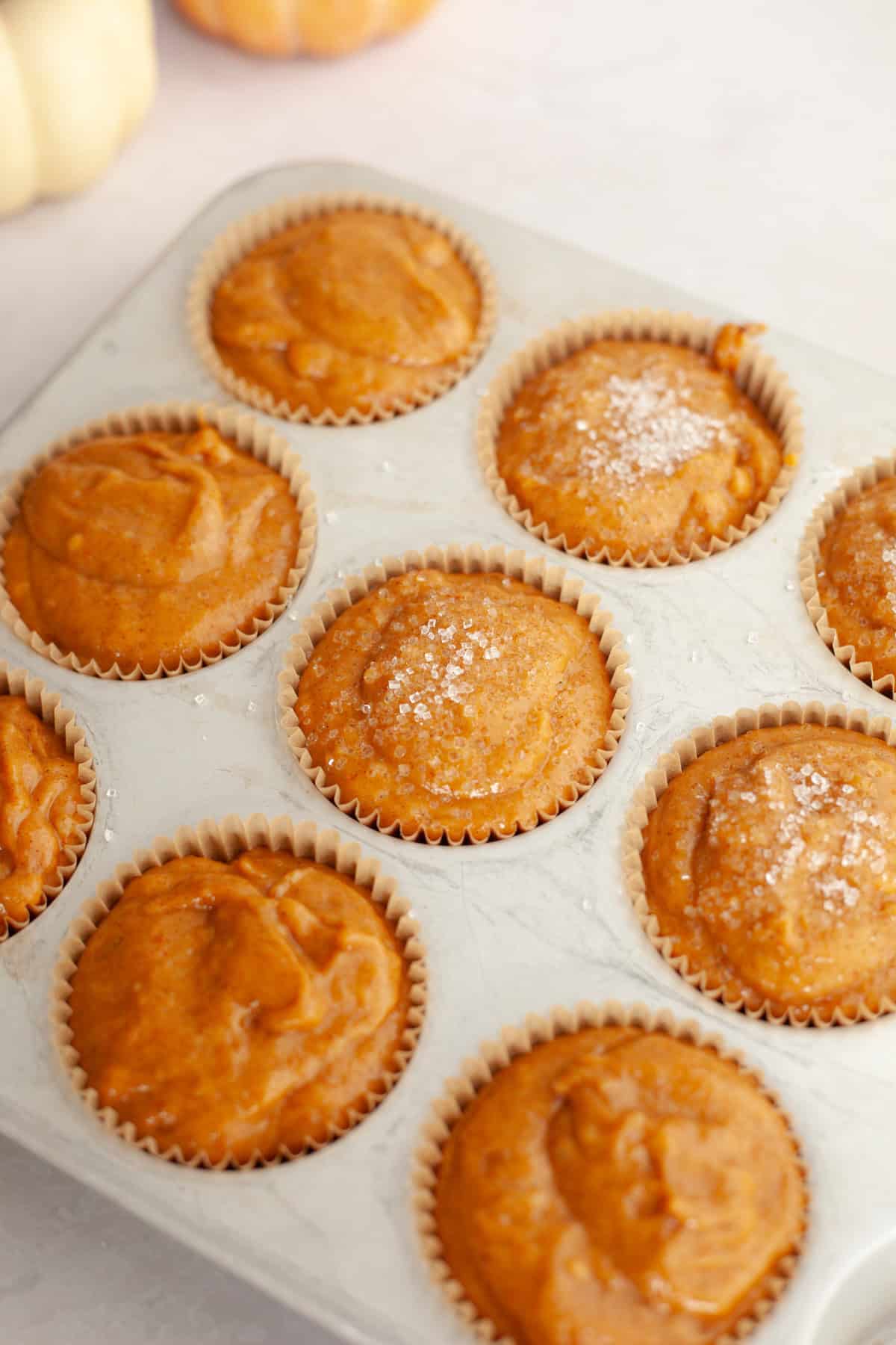 Unbaked pumpkin muffins in a muffin pan, some are topped with sparkling sugar.