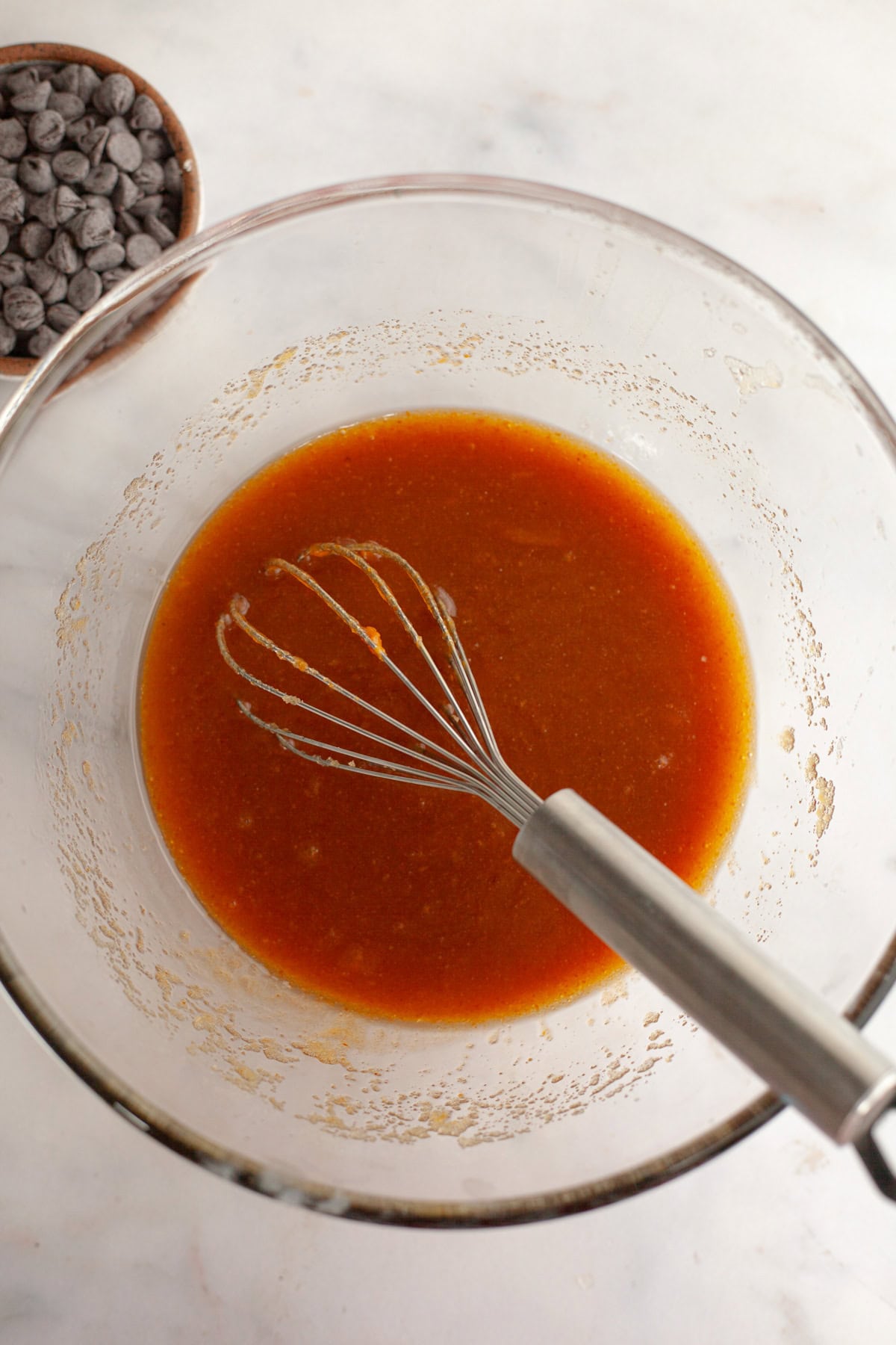 A glass bowl with a whisk and a dark orange mixture in it.