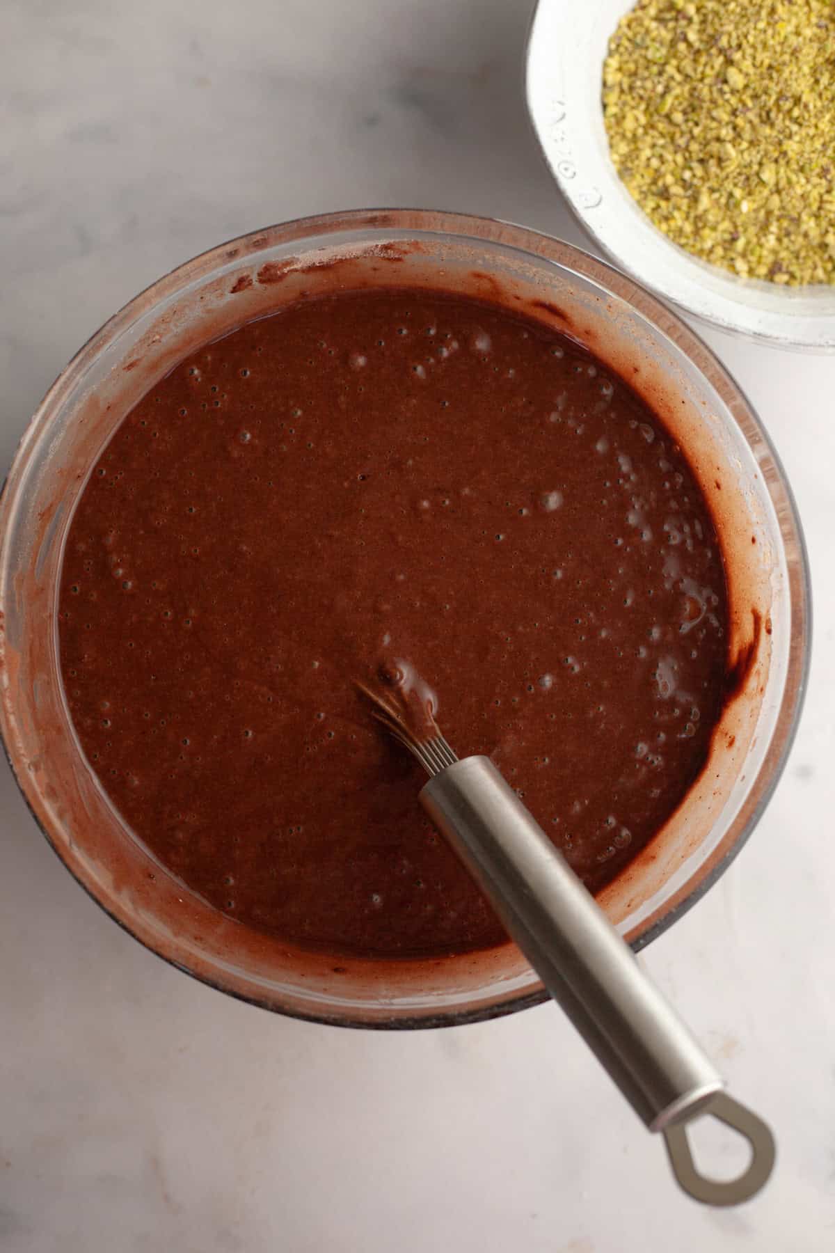 Chocolate cake batter with a silver whisk in a glass bowl.