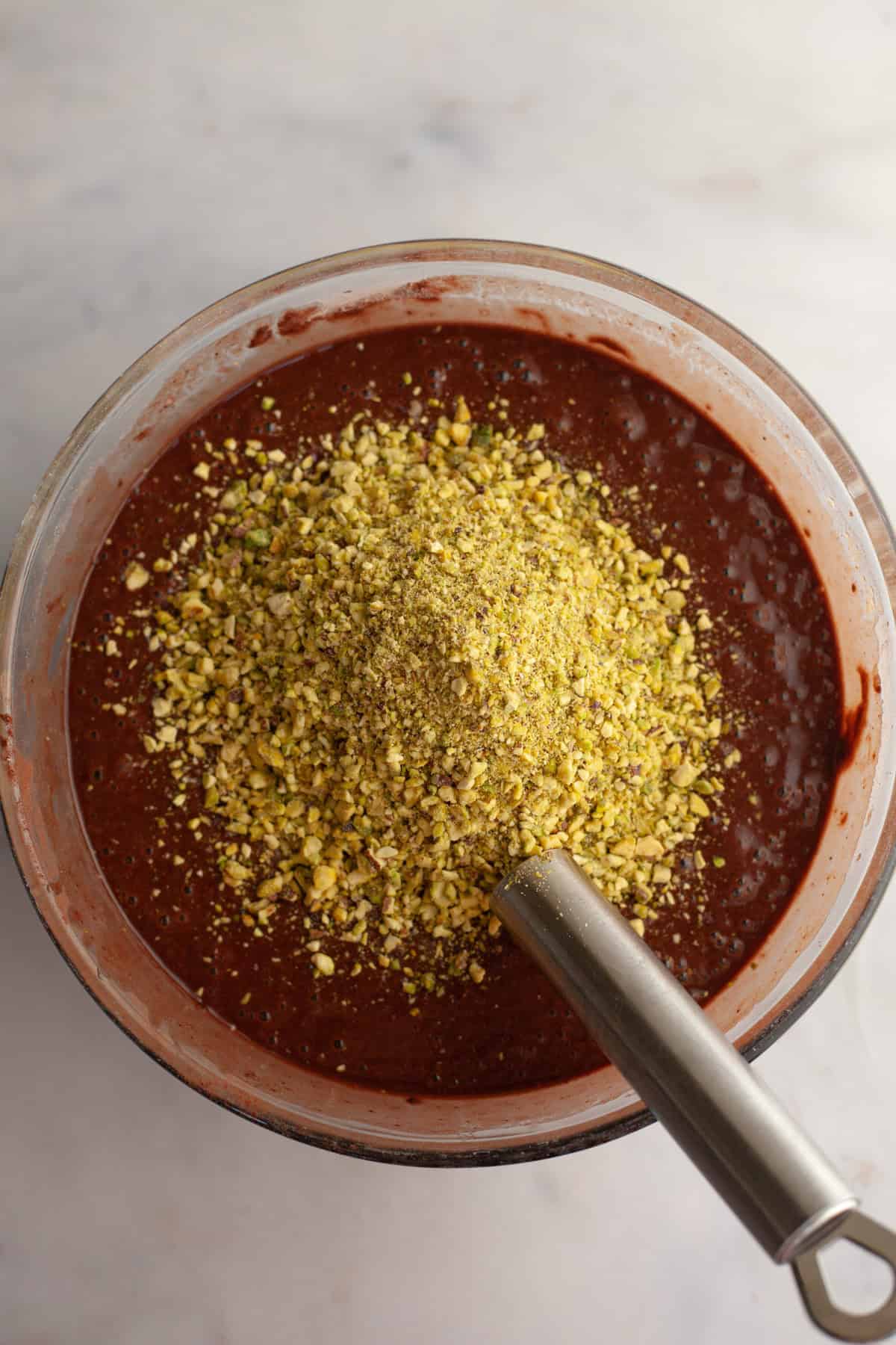 Chocolate cake batter with crushed pistachios, with a silver whisk in a glass bowl.