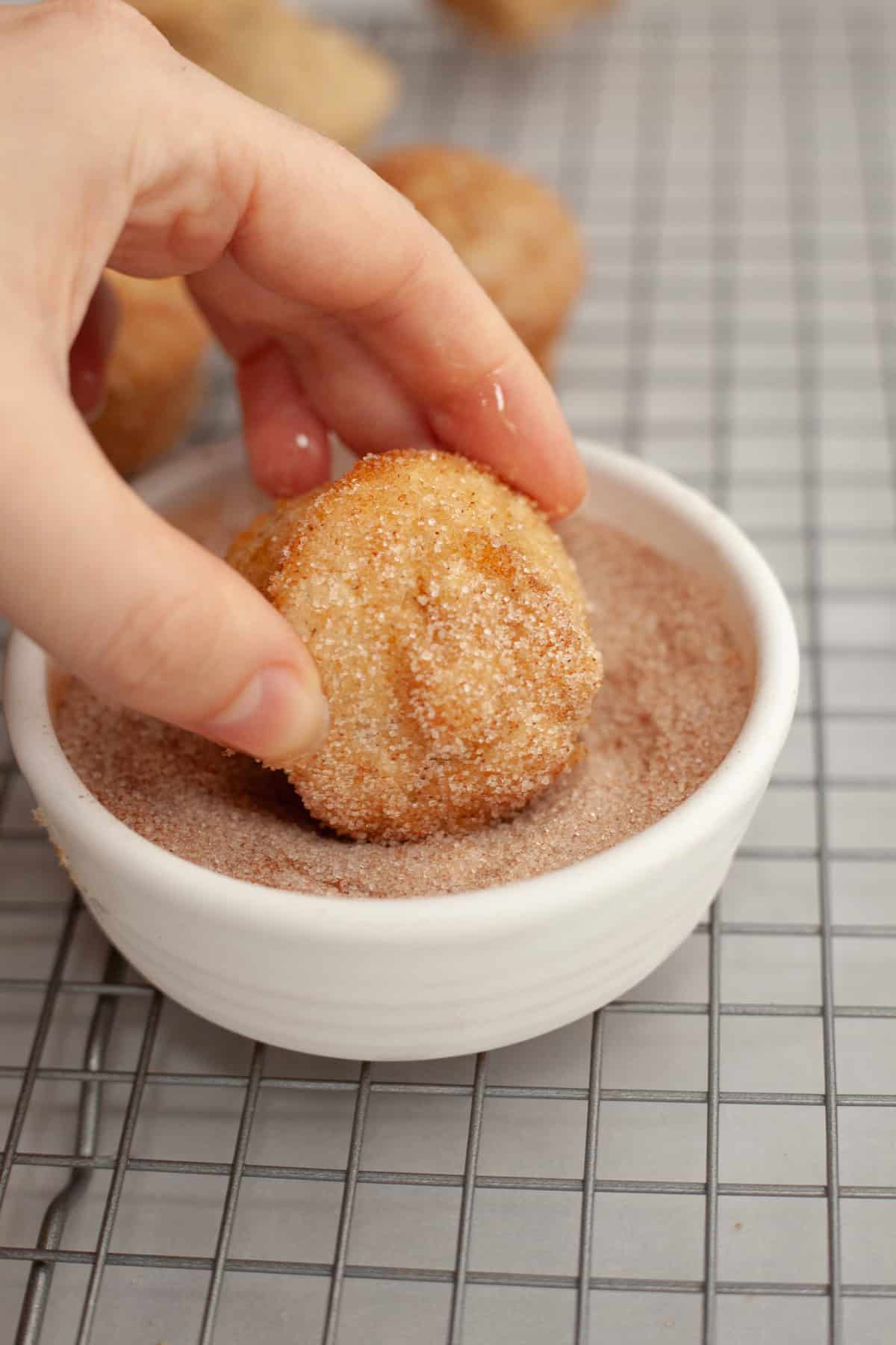 A hand rolling a mini muffin in cinnamon sugar.