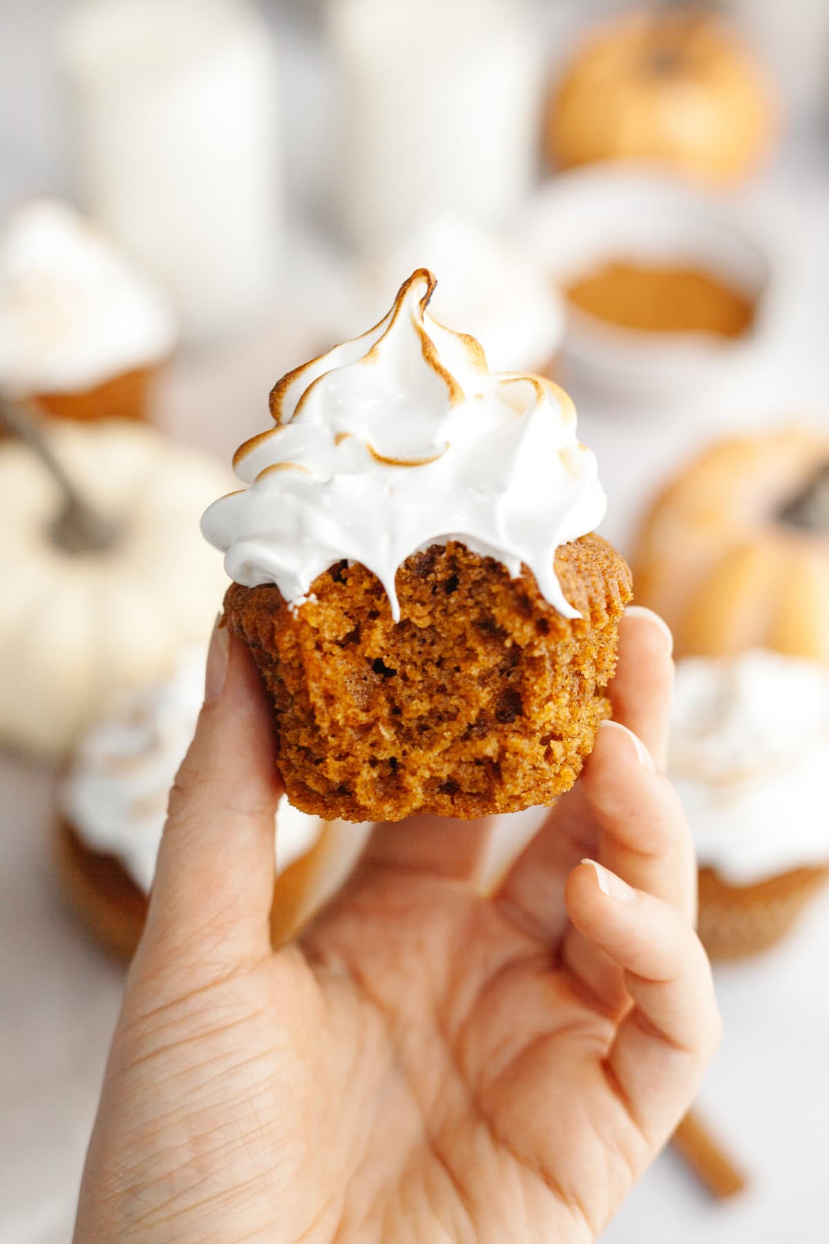 A hand holding a marshmallow frosted pumpkin cupcake with a bite taken out of it.