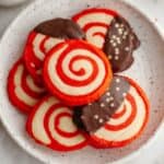 Red and white pinwheel candy cane shortbread cookies on a small plate, some of the cookies have been half dipped in chocolate.