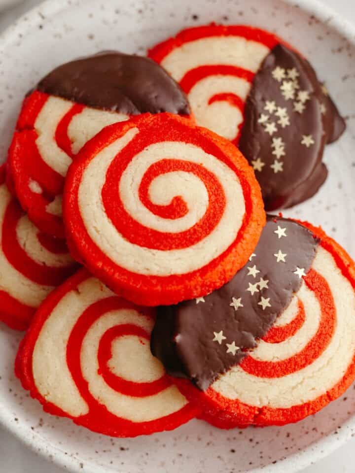 Red and white pinwheel candy cane shortbread cookies on a small plate, some of the cookies have been half dipped in chocolate.