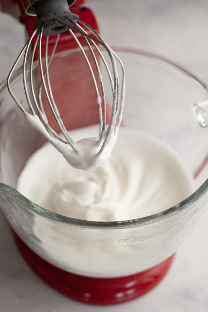 A red stand mixer with a glass bowl with meringue in it.