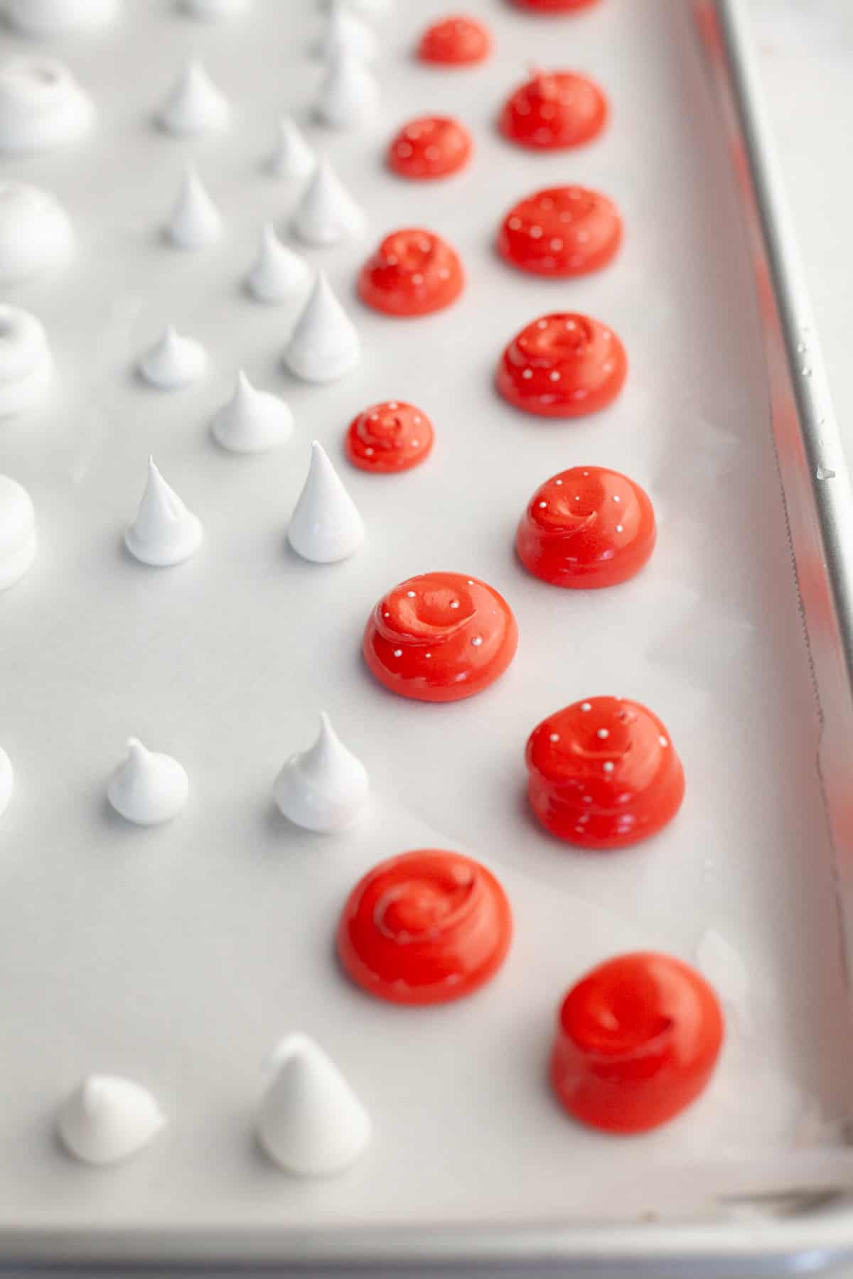 A parchment lined baking sheet with piped out red meringue mushroom tops and white stalks.